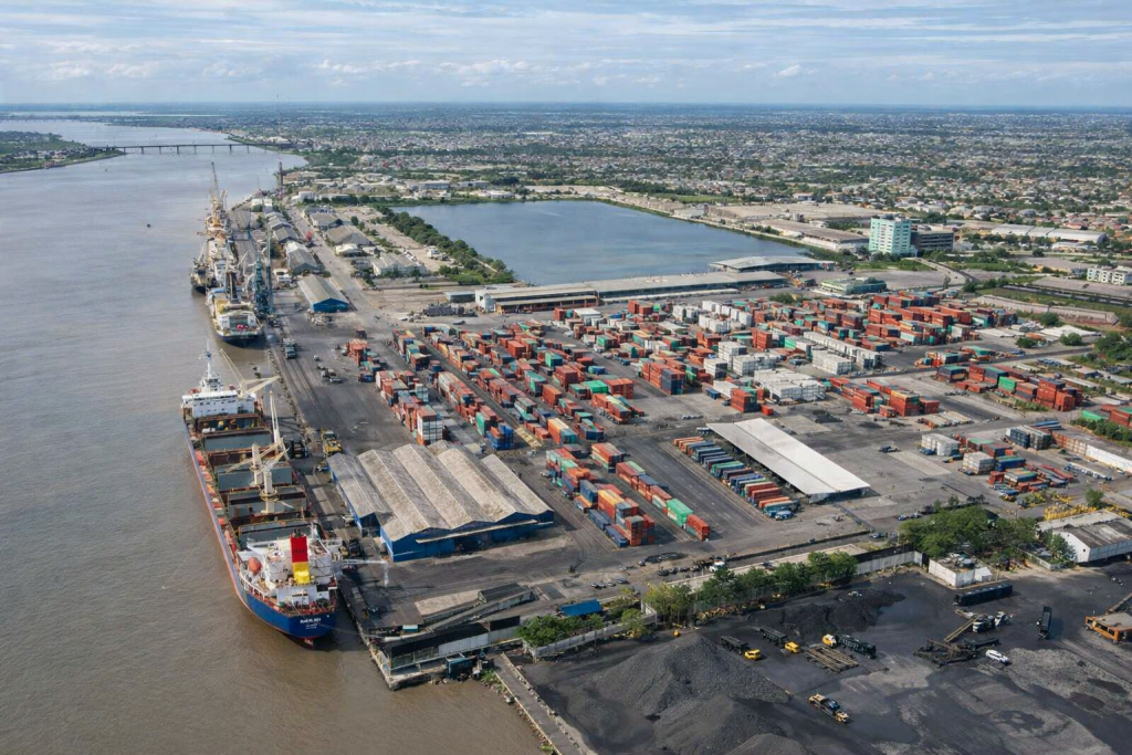 Vista aérea de un puerto industrial con contenedores y buques de carga junto a una ciudad costera.