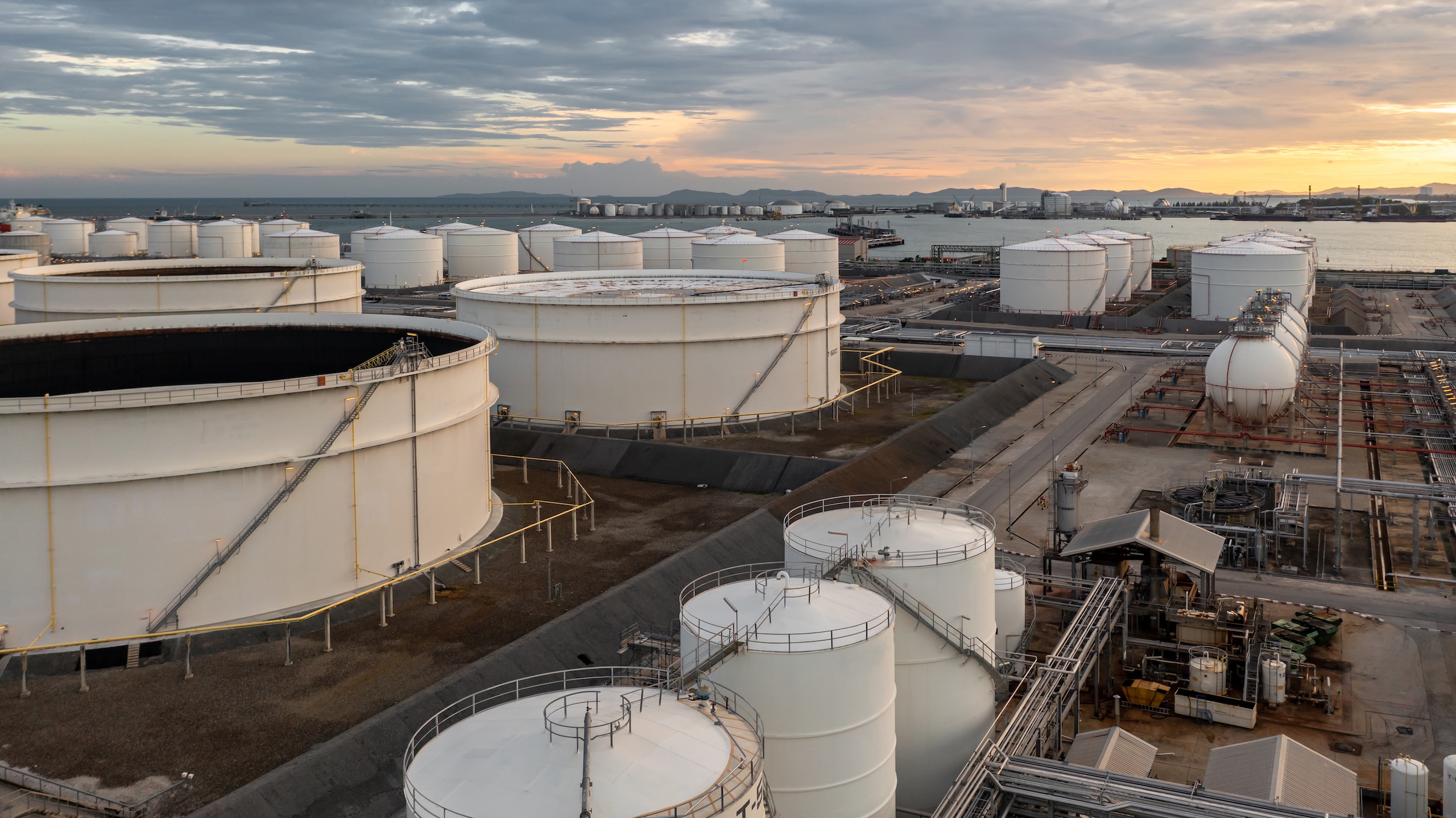 Vista aérea de una planta industrial con tanques de almacenamiento de combustibles junto a un puerto marítimo.